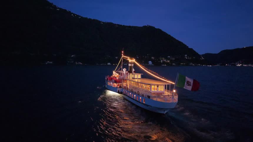 Aerial view of a brightly lit vintage ferry with an Italian flag cruising on a serene lake surrounded by dark mountains at dusk