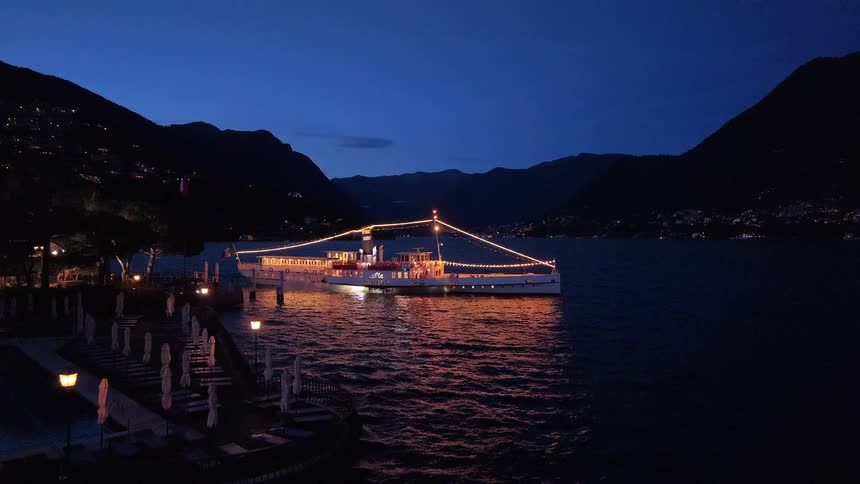 An elegant, illuminated passenger ship departs from a lakeside resort and cruises across a serene lake surrounded by mountains at night