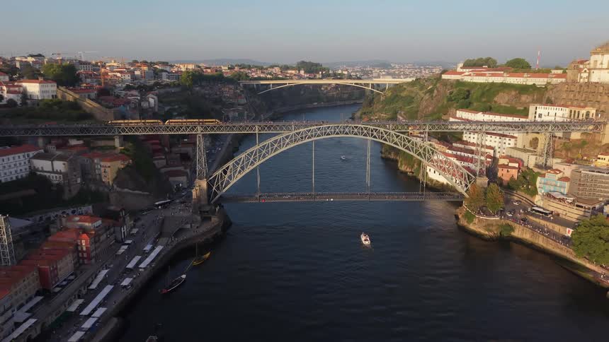 An aerial view of the Dom Luis I bridge in Porto, Portugal, as a train passes over the top and boats float on the river below at sunset