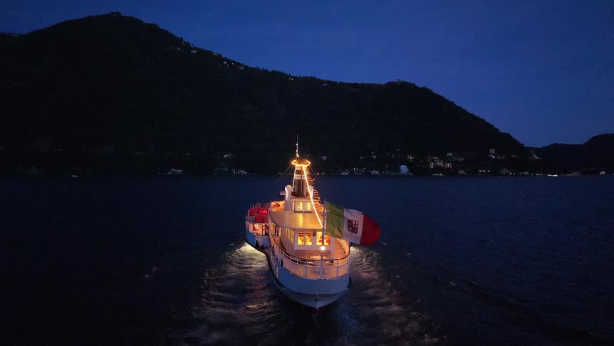 A beautifully illuminated tourist ship displaying the Italian flag cruises on a serene lake at night