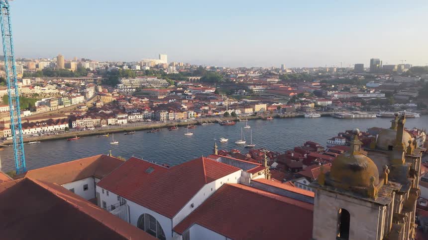 An aerial view flies over the historic rooftops and Igreja de Sao Lourenco in Porto, Portugal, revealing the Douro River with boats sailing in the golden light