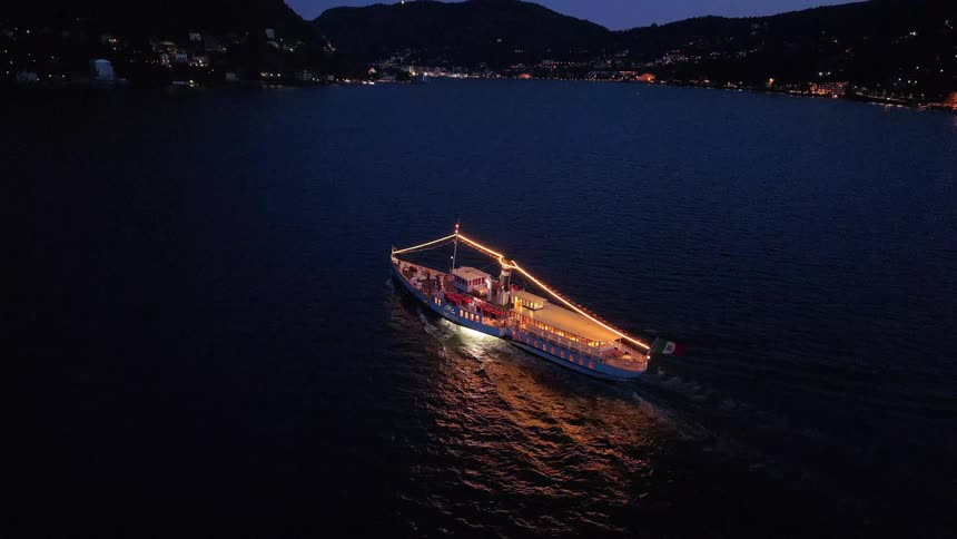 Aerial view of a classic Italian ferry, elegantly illuminated with festive lights, cruising across a serene lake at night