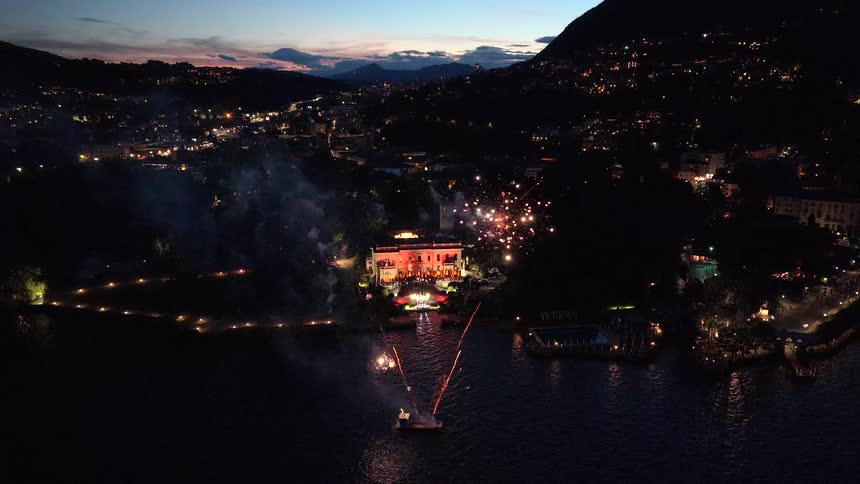 An aerial view captures a stunning fireworks display launched from a boat over a lake in front of an illuminated lakeside villa at night