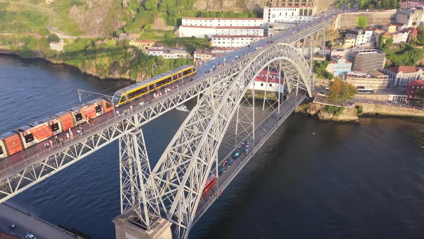 An aerial view of a metro train crossing the iconic Dom Luís I Bridge in Porto, Portugal, alongside pedestrians and above the Douro River