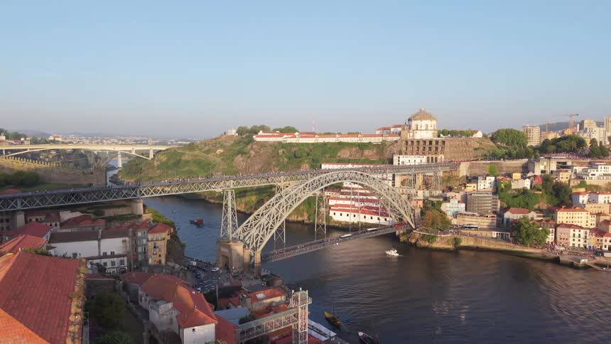 A drone flies past a construction crane to reveal a stunning view of the Dom Luís I Bridge spanning the Douro River in Porto, Portugal, at golden hour