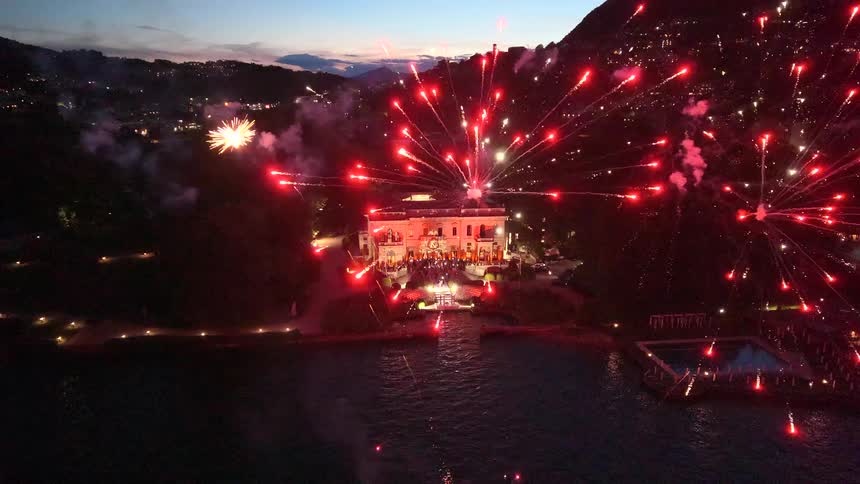 An incredible aerial view of a spectacular fireworks display lighting up the night sky over a luxurious lakeside villa during a grand celebration