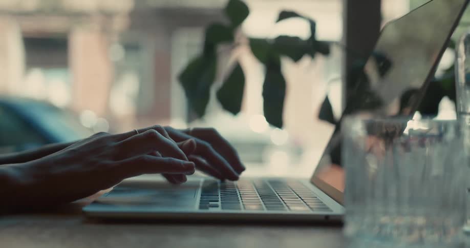 A close-up shot of a person's hands typing on a laptop in a cafe with a soft, natural light from a nearby window