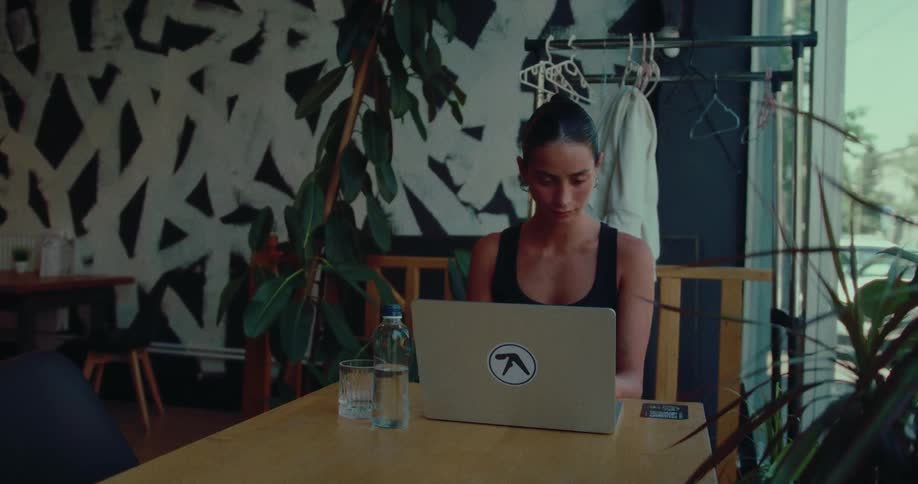 A young woman works on her laptop at a table in a stylish, plant-filled cafe