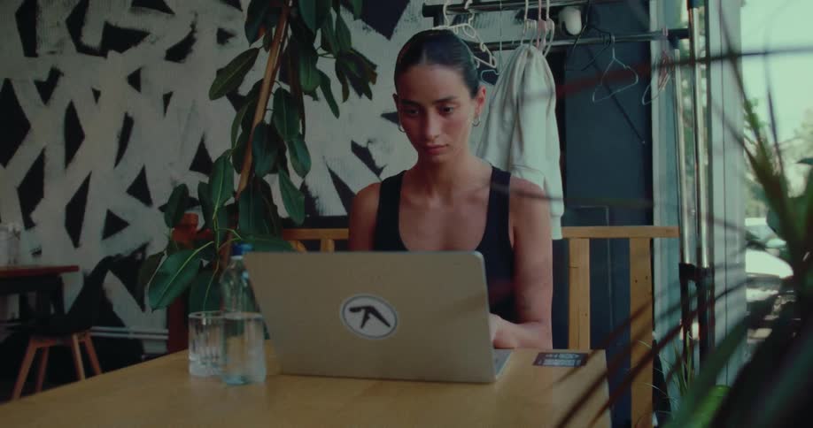 A young woman opens her laptop to work at a table in a modern, plant-filled cafe by a large window
