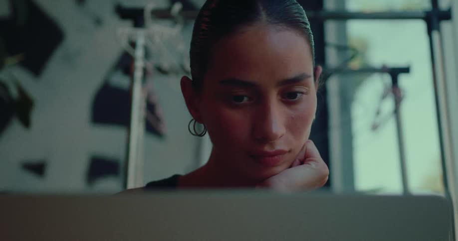 A focused young woman with her hair pulled back works intently on her laptop in a modern, softly lit space