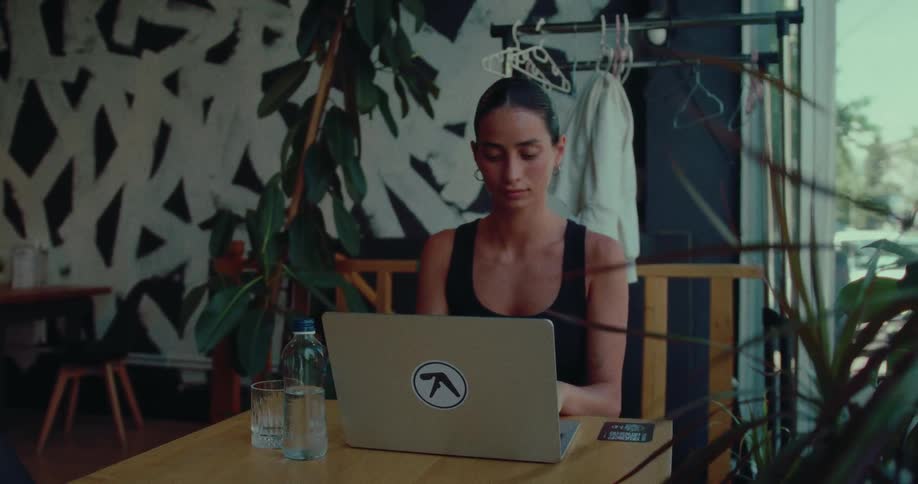 A focused woman works on her laptop at a table in a stylish, plant-filled cafe