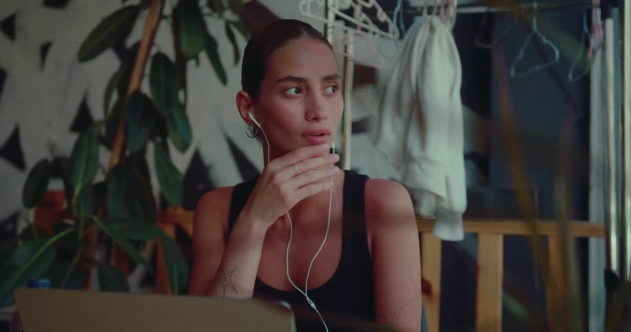 A pensive young woman with earphones listens to music while looking at her laptop in a sunlit, plant-filled room