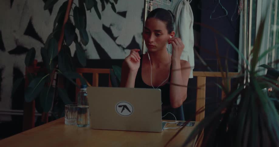 A focused young woman puts in earphones and begins working on her laptop in a dimly lit, plant-filled space