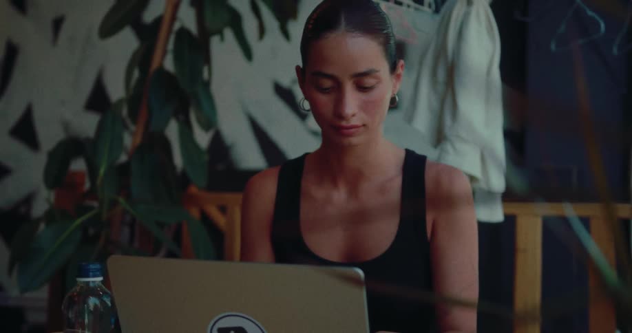 A focused young woman in a black tank top works on her laptop in a modern, plant-filled space