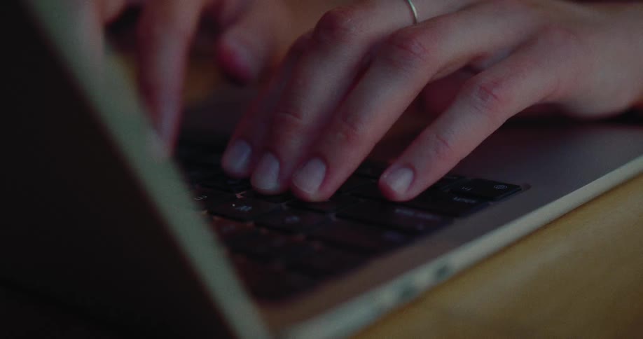 A person opens their laptop and begins typing on the keyboard in a dimly lit setting