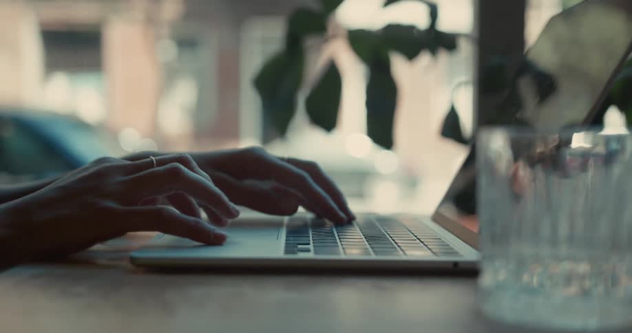A side profile shot of a person opening a laptop on a wooden table and beginning to type in a brightly lit cafe