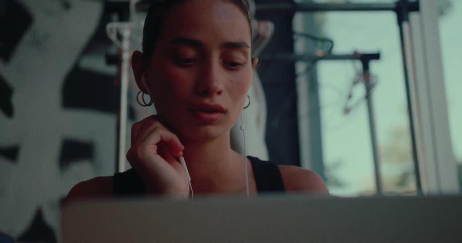 A focused young woman listens to music on earbuds while working on a laptop in a modern creative space
