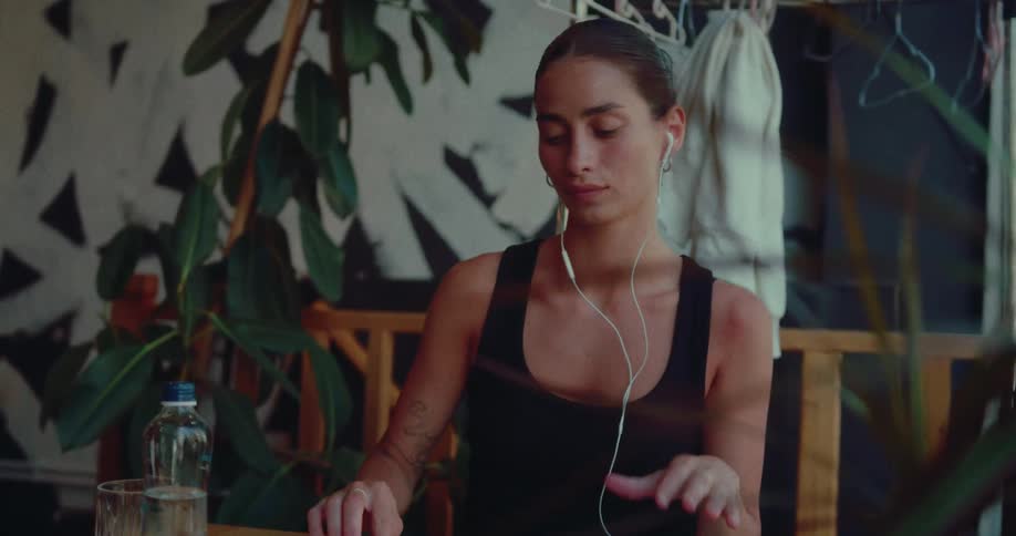 A young woman with tattoos finishes working remotely on her laptop, closes it, and removes her earbuds in a plant-filled cafe