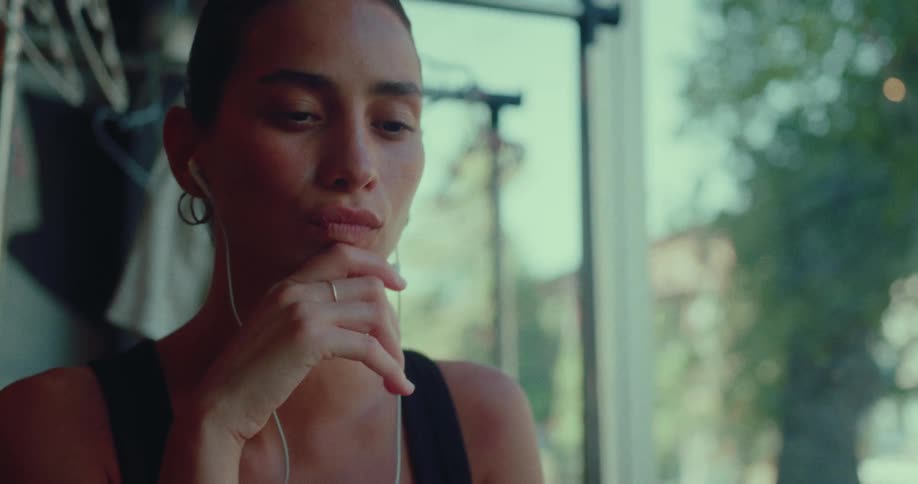 A young woman wearing earbuds works on her laptop, looking pensive as she sits by a sunlit window
