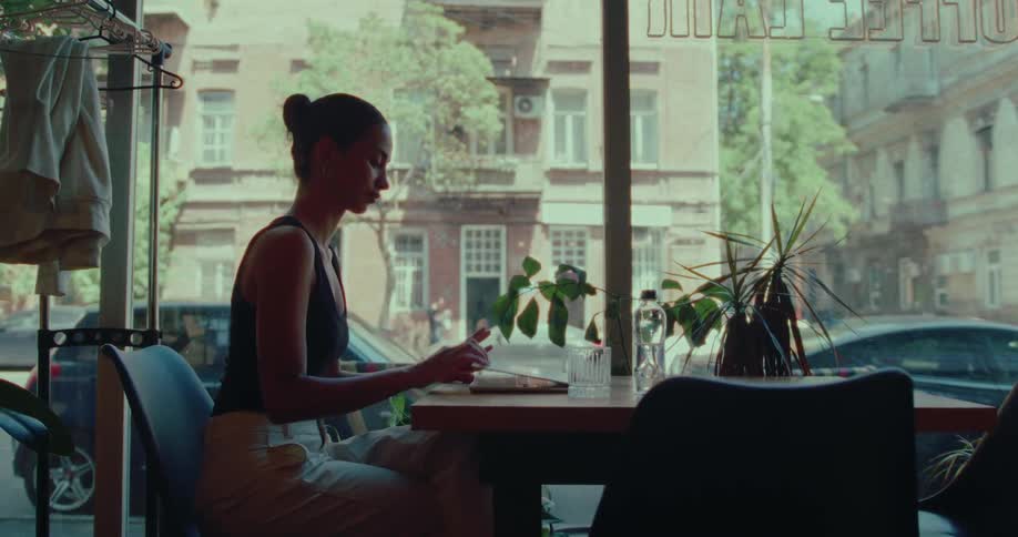 A contemplative young woman pauses from her work in a stylish city cafe to watch the world go by outside the window