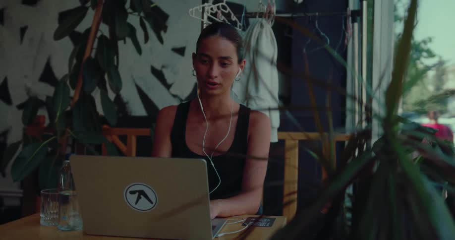 A focused young woman wearing earphones works remotely on her laptop, engaging in a video call from a stylish cafe patio