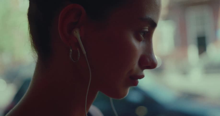 Close up profile shot of a young woman with hoop earrings listening to music on her wired headphones while sitting by a window in the city