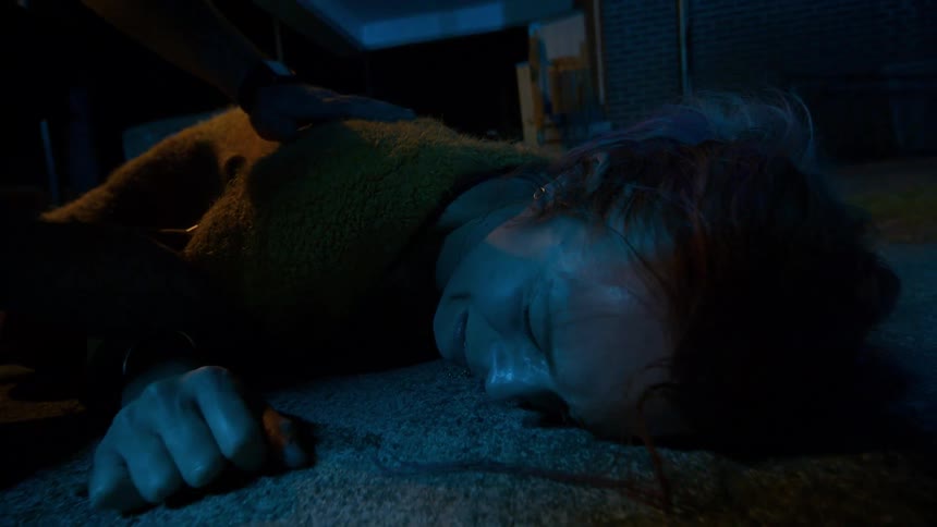 A distraught woman lies on the ground in pain at night while a first responder tends to her under flashing blue lights