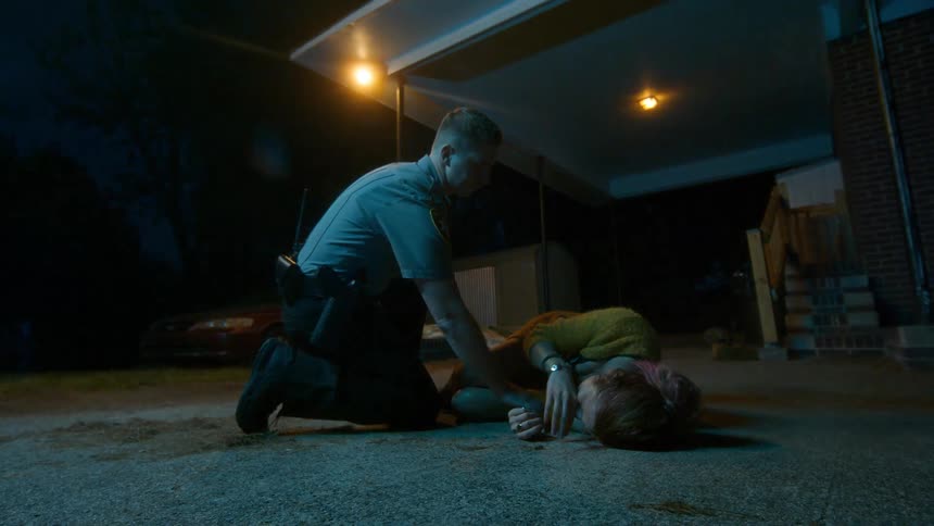 A police officer kneels beside a wounded woman lying on the ground at night, checking her pulse