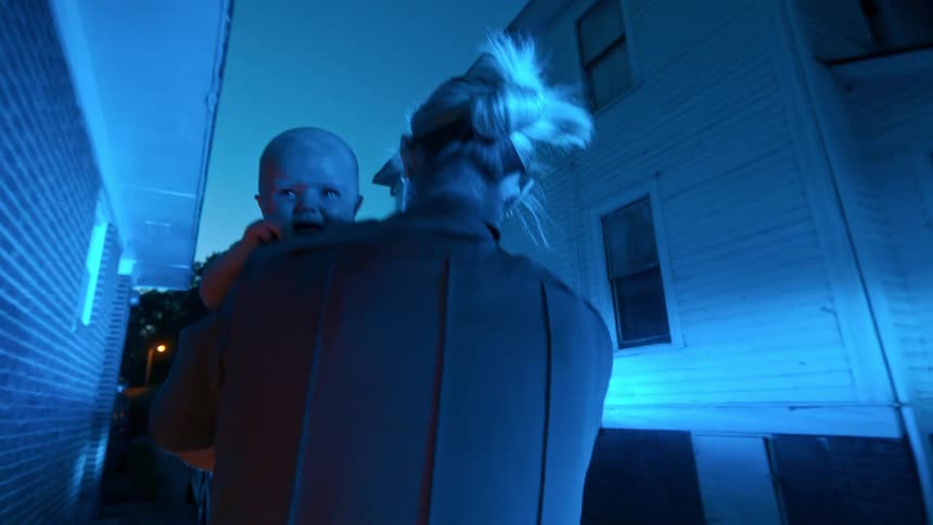 A female police officer holds a crying baby at night as flashing blue lights from her patrol car illuminate the dramatic scene