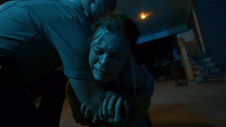 A police officer restrains a crying and hysterical woman during a chaotic nighttime arrest under flashing lights