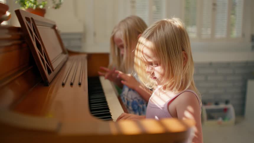 Adorable blonde sisters sit side-by-side, their small hands exploring the keys of a wooden piano in warm morning light
