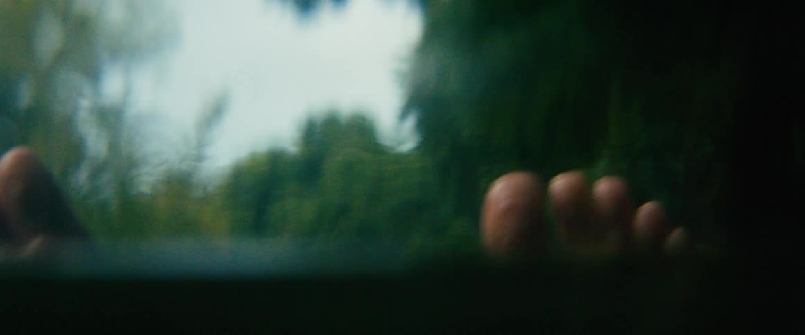 A close-up shot shows a person's bare feet resting on the car window during a moody road trip through a blurry green countryside