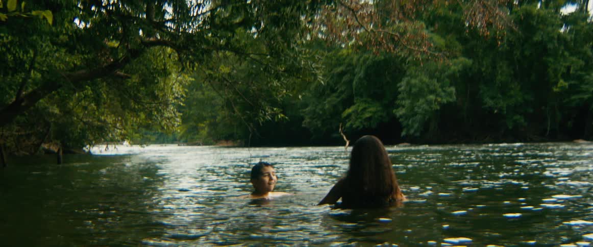 A woman and a young boy relax in a serene, tree-lined river, and the boy happily floats on his back