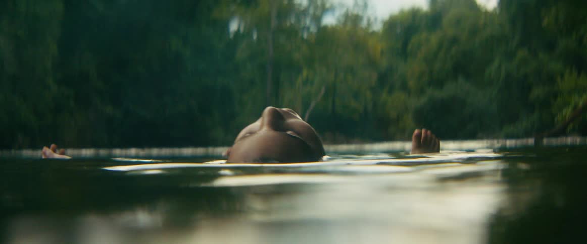 A serene person floats peacefully on their back in a calm river surrounded by a lush forest as the light fades to darkness