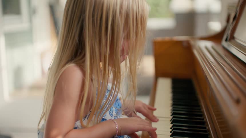 An adorable young girl with long blonde hair carefully presses the keys of a wooden piano in a sunlit room