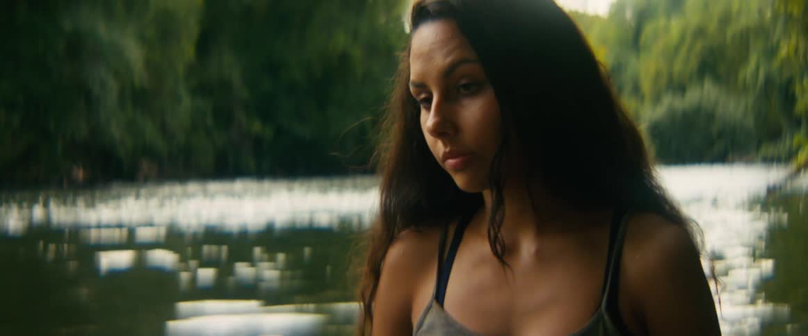 A pensive young woman's expression shifts from sadness to a gentle smile as a sudden rain shower begins by a tranquil river