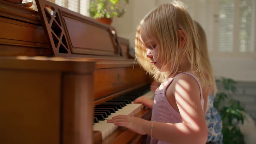 Two adorable young blonde sisters playfully press the keys of a wooden piano together in a beautifully sunlit room