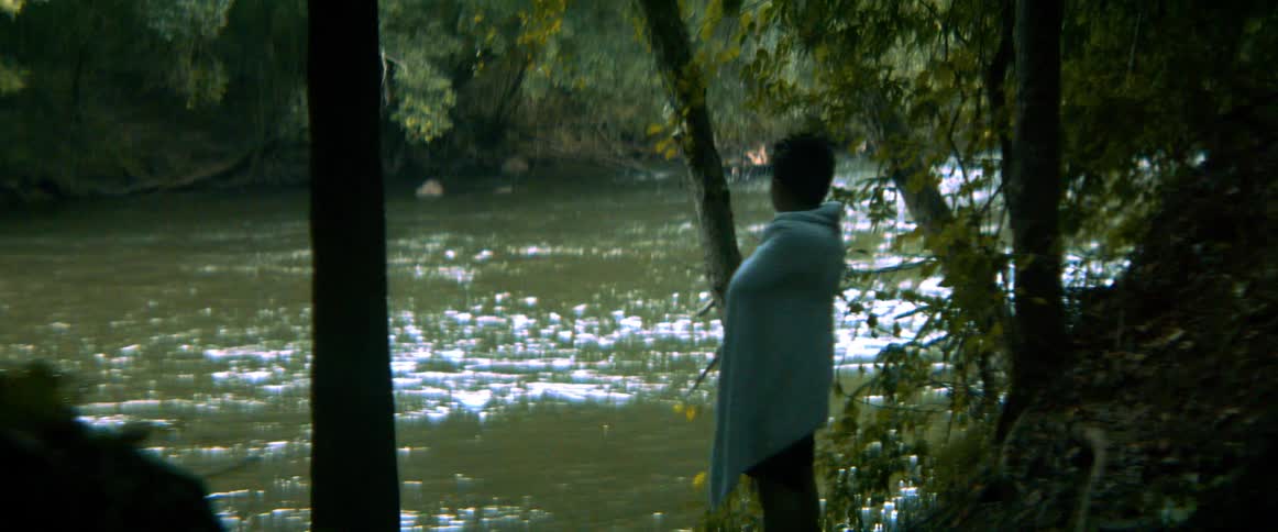 A young boy in silhouette wraps himself in a towel while standing on the bank of a shimmering river