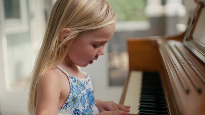 A cute young blonde girl happily sings and concentrates while playing an upright wooden piano in a sunlit room