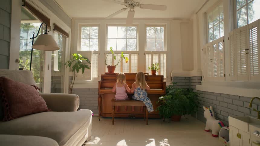 Two young sisters sit side-by-side playing an upright piano in a bright and airy sunroom as the camera slowly pulls back to reveal the cozy space