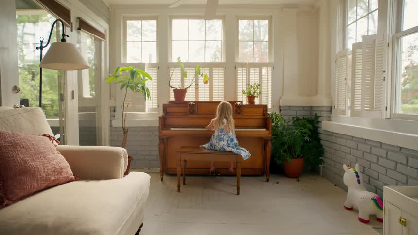 A little girl in a blue and white dress plays an old upright piano in a bright and cozy sunroom