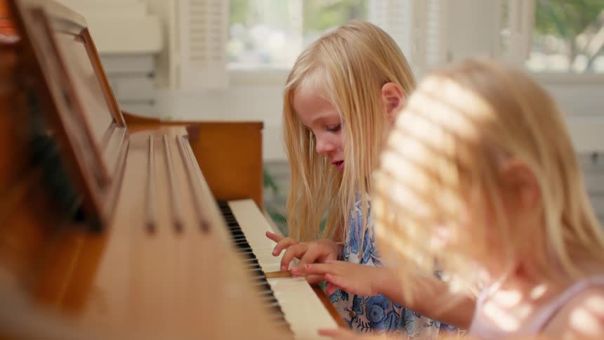 Two adorable blonde sisters playfully learn to play the piano together in a sunlit room