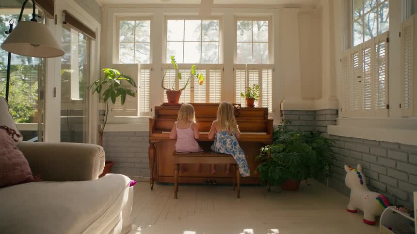 Two young sisters sit side-by-side playing a duet on an old upright piano in a bright and airy sunroom at home