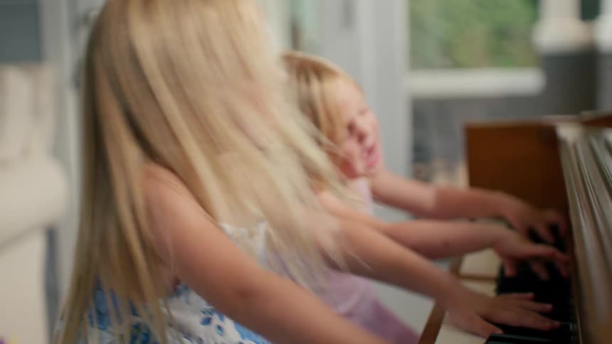 Two energetic young blonde sisters have a blast singing and playing the piano together at home