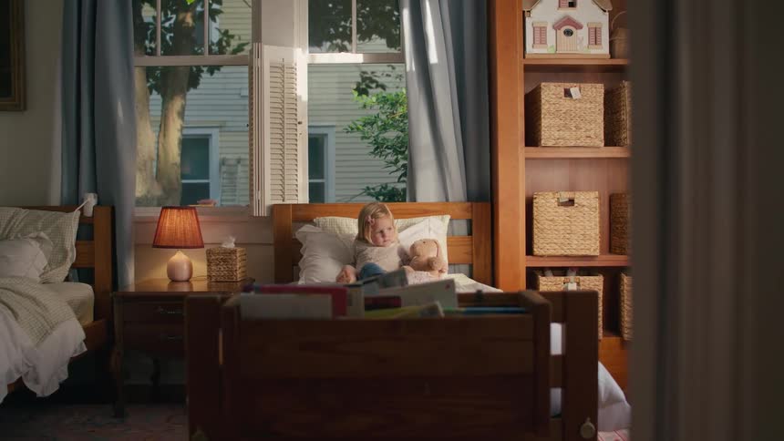 A sad-looking little girl sits up in her bed with her doll in a sunlit bedroom