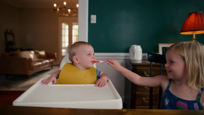 A young girl smiles as she carefully feeds her baby brother in his high chair at the dining room table