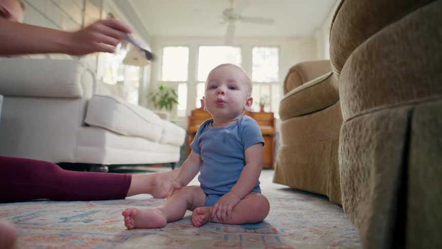 Low-angle shot of a mother feeding her infant son with a spoon as they sit together on the living room rug