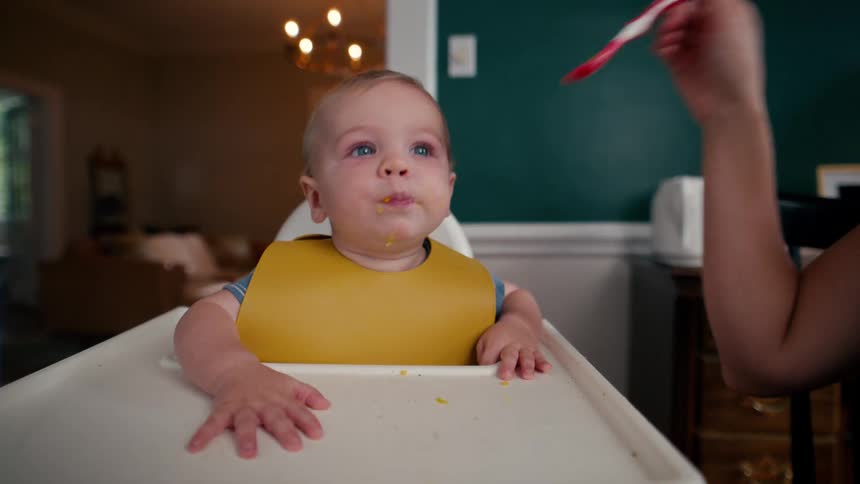 A mother spoon-feeding her cute baby boy wearing a yellow bib in his high chair