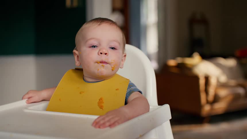 A cute and messy baby with blue eyes is being spoon-fed in a highchair