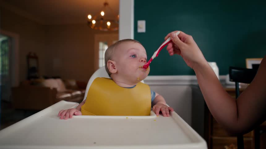A mother spoon-feeds her sleepy baby boy puree in his high chair at the dining room table